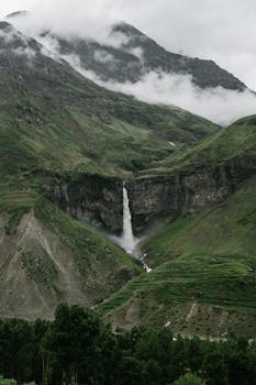 Breathtaking waterfall cascading down a lush green mountainside surrounded by mist and clouds.