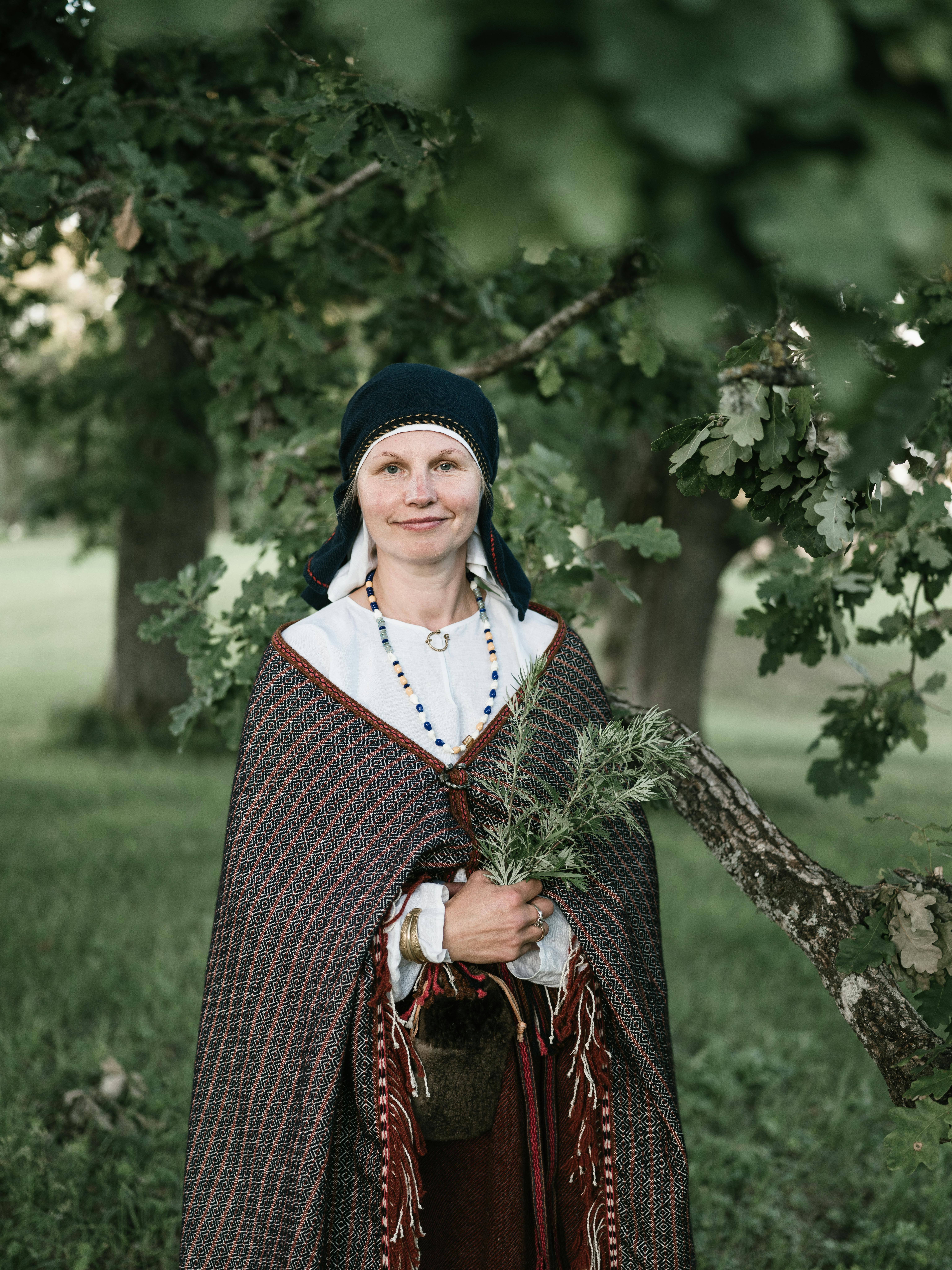 A woman in traditional attire holds wild herbs, standing in a lush outdoor setting.