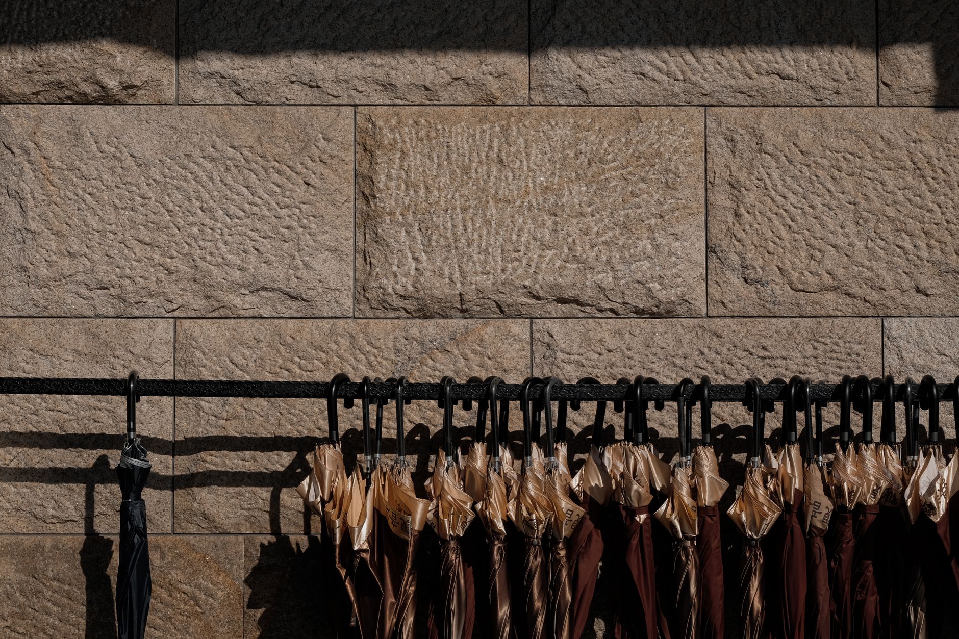 A close-up of umbrellas hanging on a black metal rack against a rough-textured concrete wall in sunlight.
