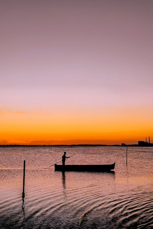 Side view of person standing in boat and floating on calm river at sunset
