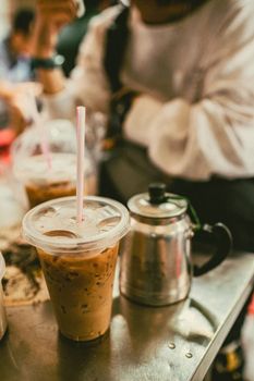 Close-up of iced coffee in a plastic cup with a straw on a metal table beside a teapot.