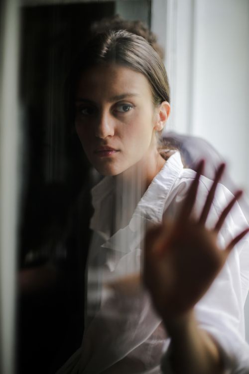 Free A woman in a white shirt gazes thoughtfully through a window, hand touching the glass. Stock Photo