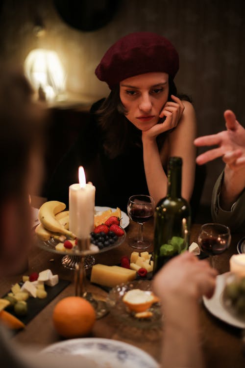 Woman in Black Tank Top Wearing Red Hat Sitting Beside Table With Bottles and Candles