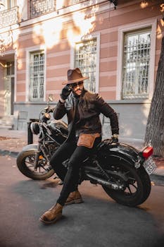 Fashionable man in leather jacket and hat sitting on a parked motorcycle on an urban street.