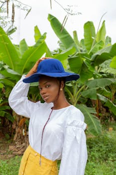 Self assured young black female in trendy outfit adjusting hat and looking at camera while relaxing in tropical park during summer holidays