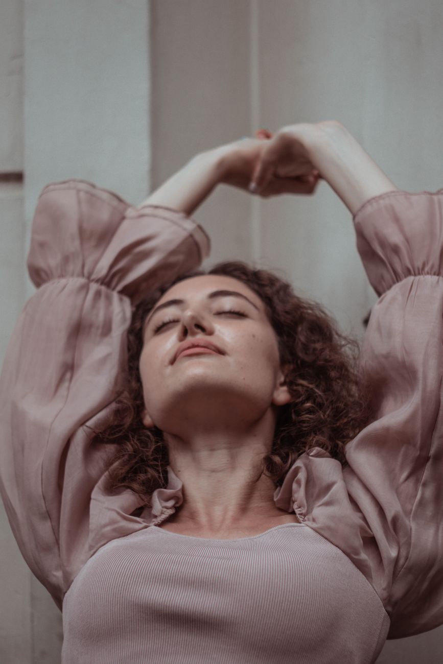 Young female with brown curly hair and eyes closed wearing stylish dress stretching arms above head
