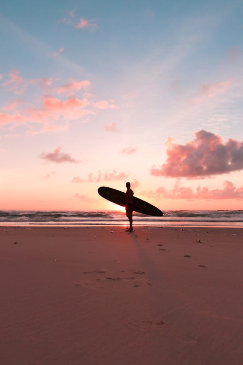 Free Silhouette of a Person Carrying a Surfboard Standing on Shore Stock Photo