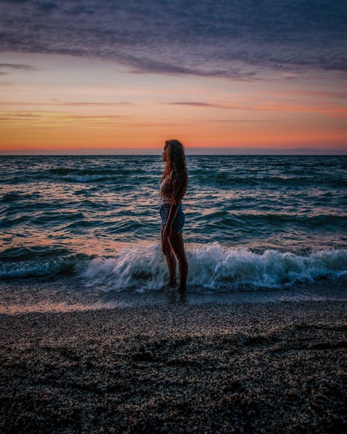 Free Full body of anonymous female standing on sandy beach near foamy wave against bright sunset sky in tropical country in evening Stock Photo