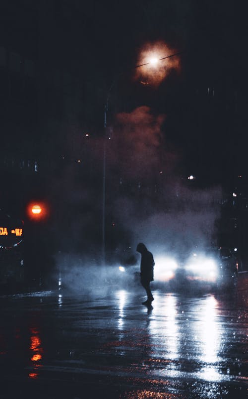 Silhouette of person walking on city street near car at night