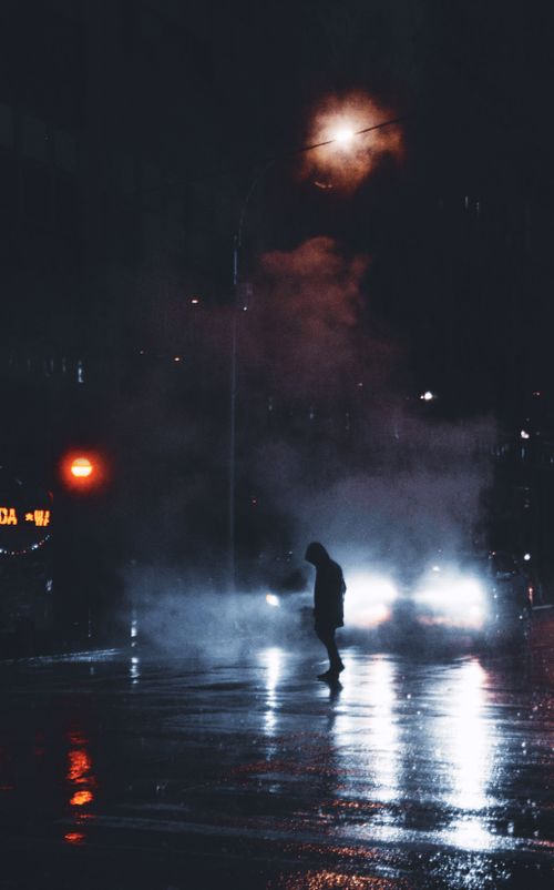 Free Side view of unrecognizable person in warm hooded outerwear crossing road near stopped vehicle with glowing headlights on rainy night Stock Photo