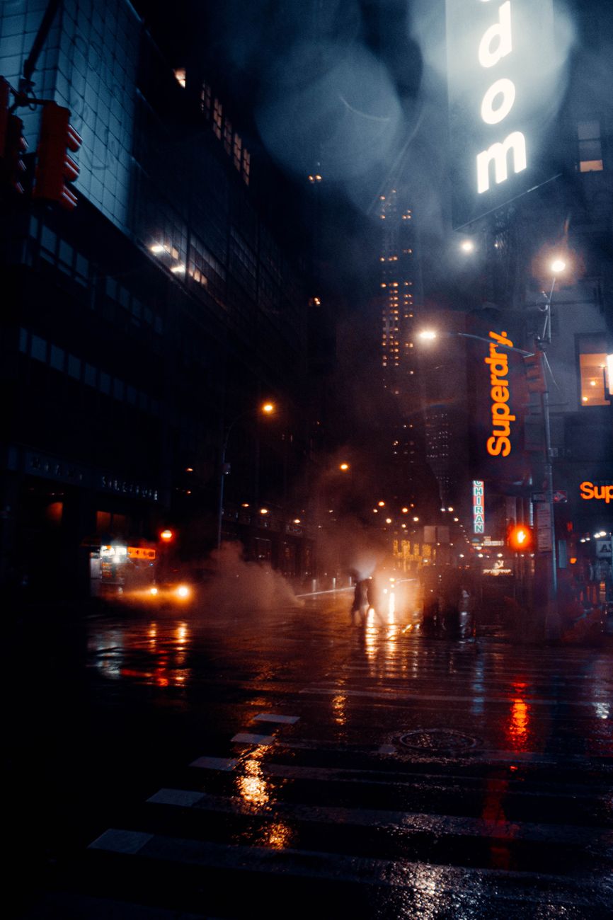 Contemporary multistage building exteriors and roadway with vehicles illuminating town in evening in rainy weather