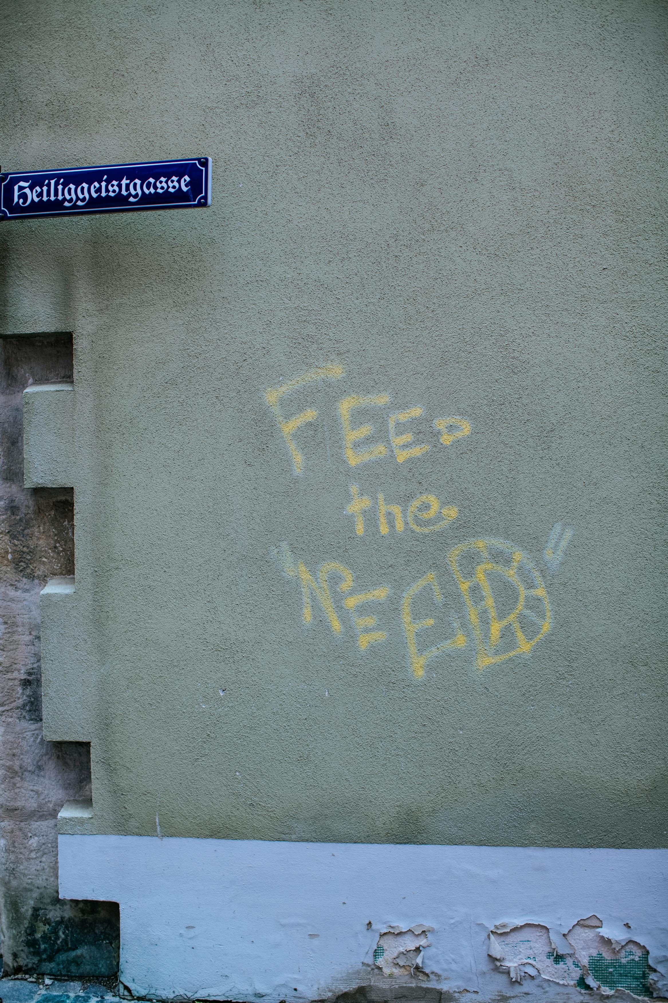 Street graffiti with slogan 'Feed the Need' on a concrete wall next to Heiliggeistgasse sign.