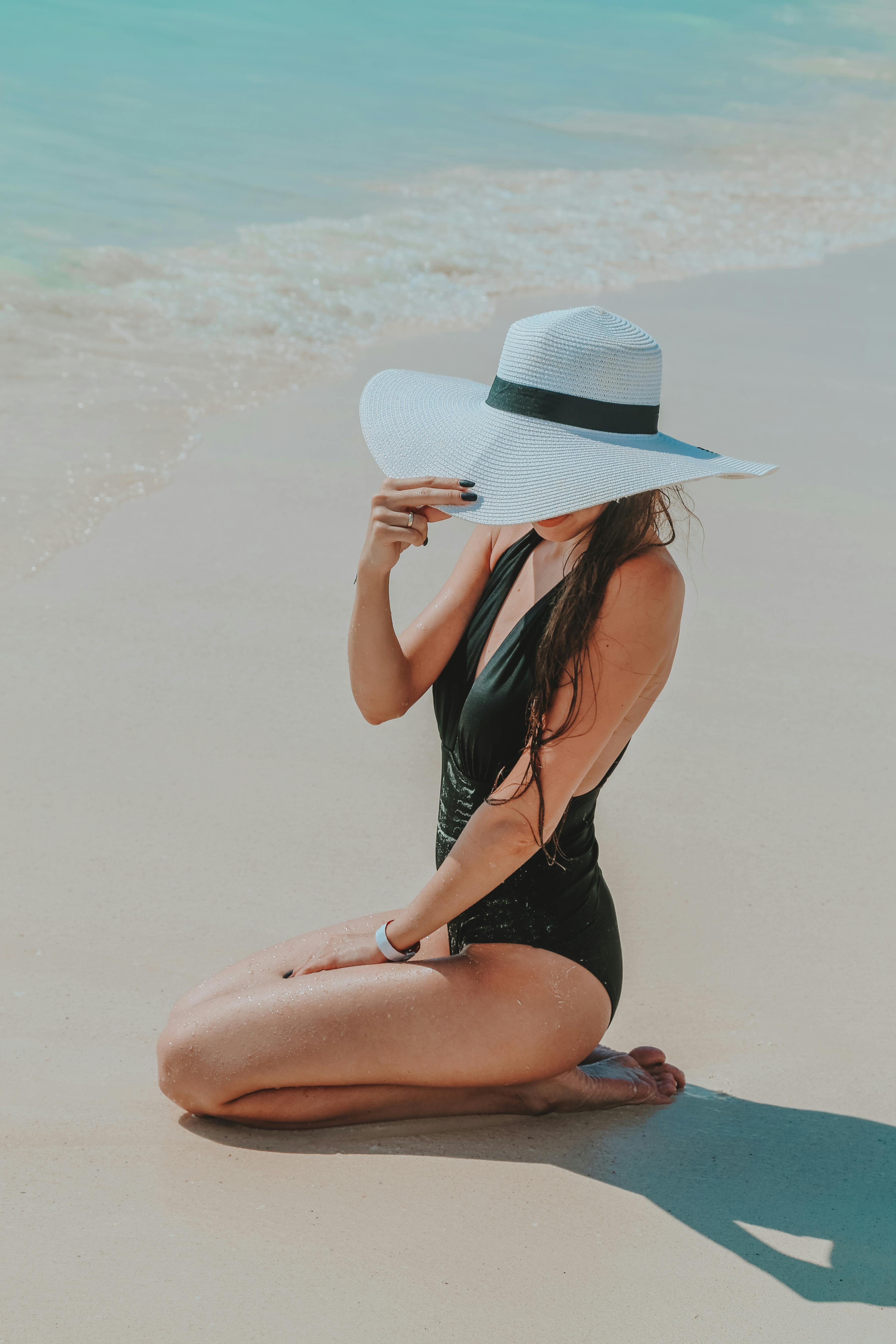 From above side view of anonymous trendy slim barefoot female tourist in black swimwear touching hat while sitting on sandy ocean beach