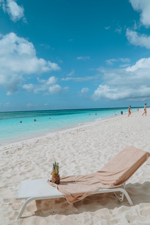 From above of sun bed with towel and ripe pineapple on sandy sea shore with unrecognizable tourists under blue cloudy sky