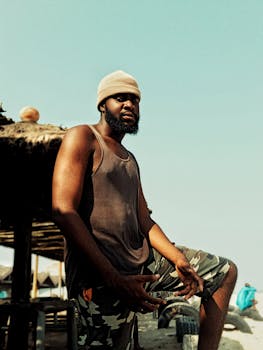 African man in casual attire relaxing on a beach in Accra with a thoughtful expression.