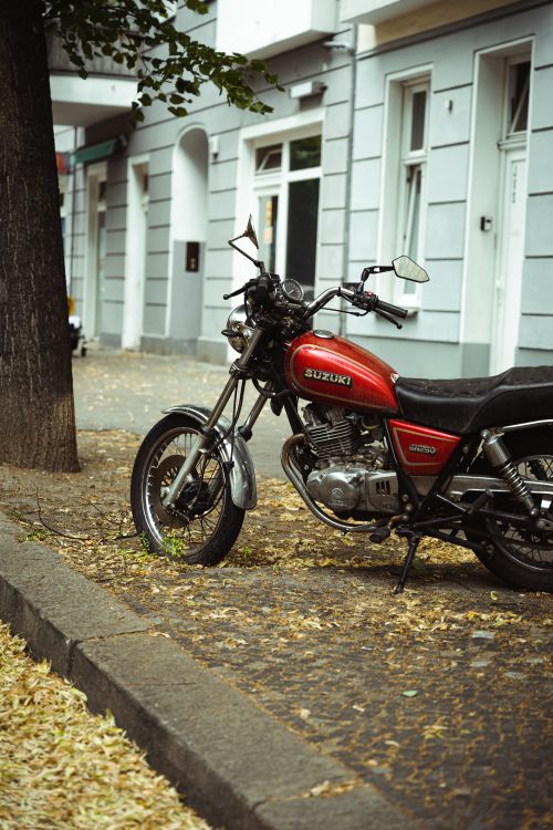 Free Red Suzuki motorcycle parked on a roadside next to a building with fallen leaves on the sidewalk. Stock Photo