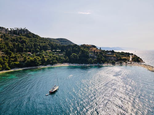 From above of sailboat sailing on rippled sea near mount with greenery trees under cloudy sky in evening