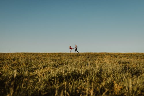 Distant happy man and woman in love holding hands and running on endless green meadow with blue sky