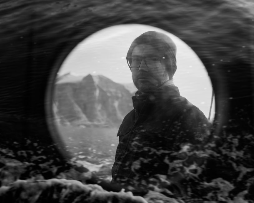 A black and white portrait of a man viewed through a glass window, capturing reflections and a moody landscape behind.