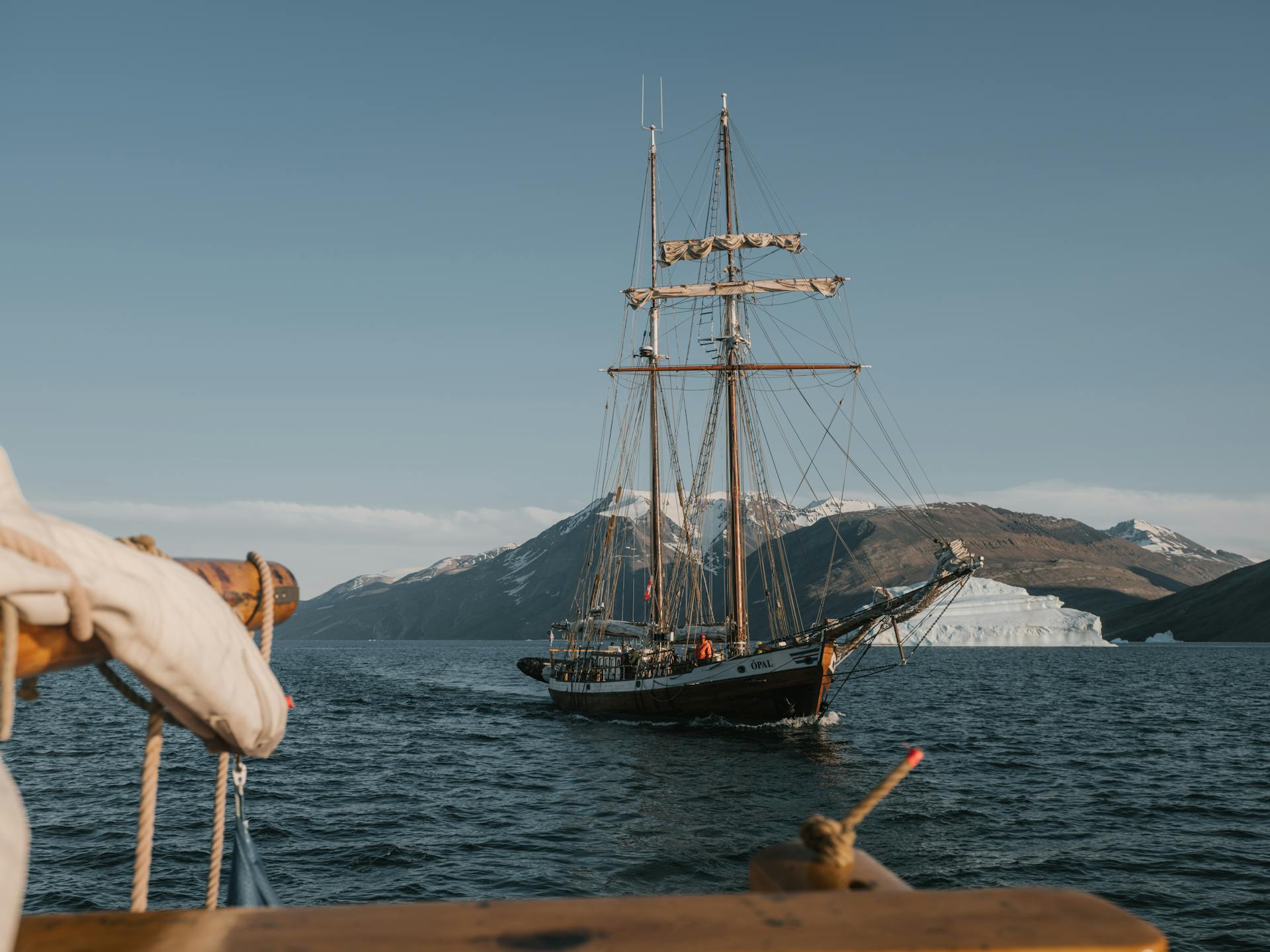 Historic sailing ship in the Arctic, mountains and iceberg in the background.