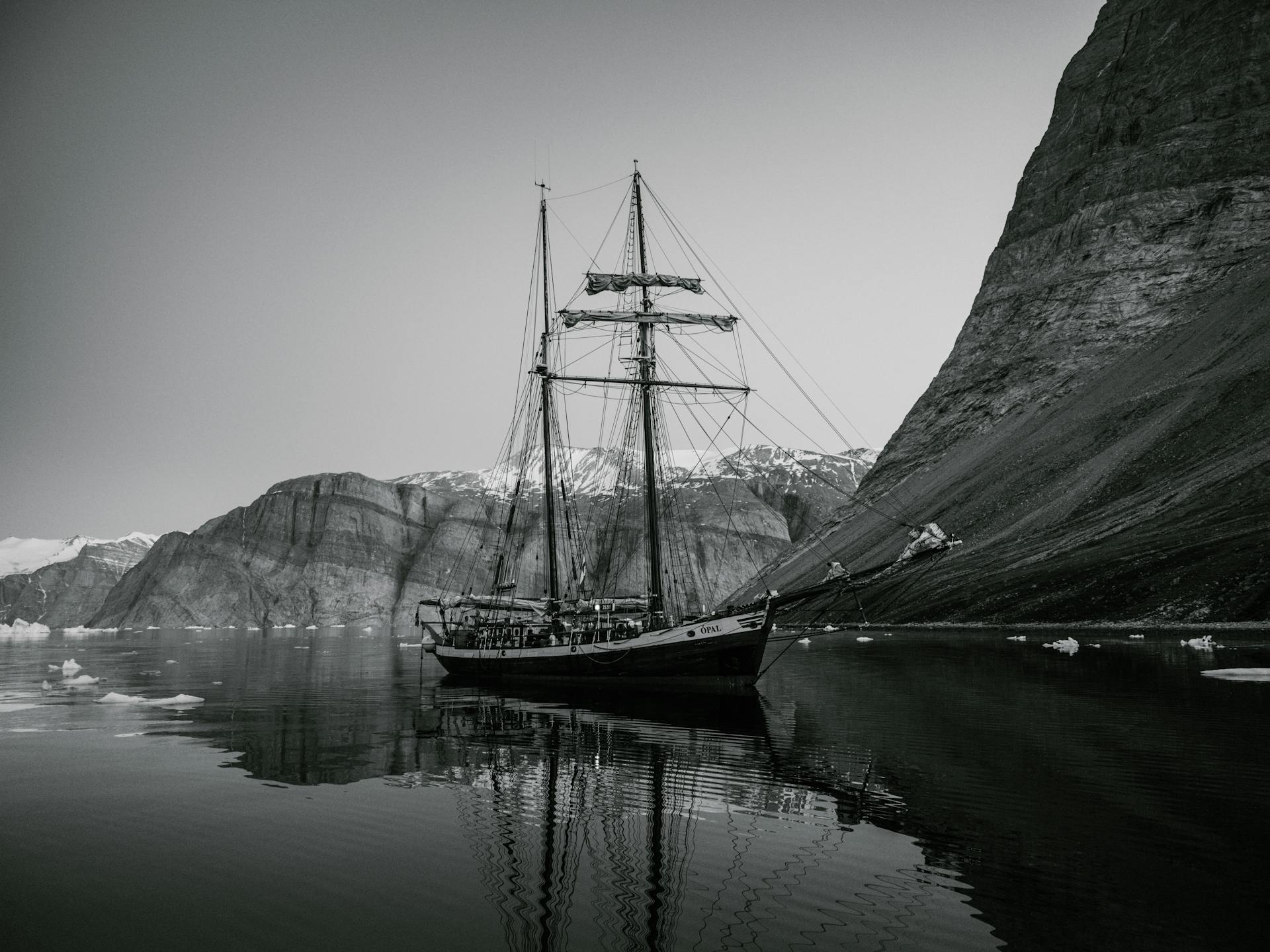 Black and white image of a schooner on calm water with rocky mountains in the background.