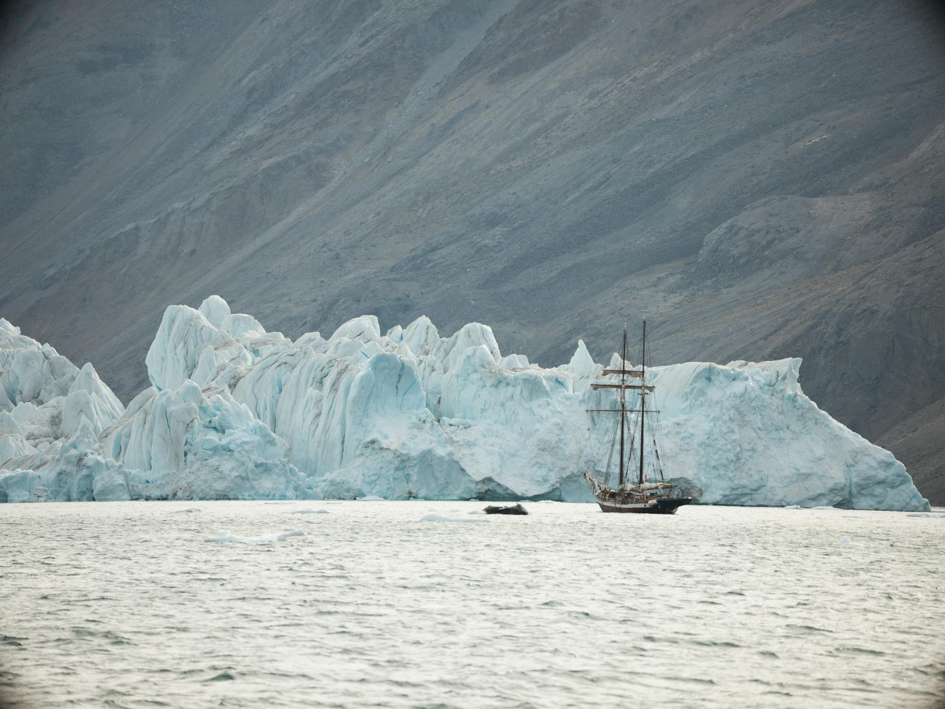 A stunning view of a schooner sailing past a towering iceberg in calm waters.