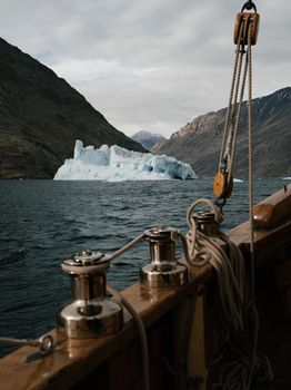 Sailboat on ocean near iceberg with rocky mountains in background.