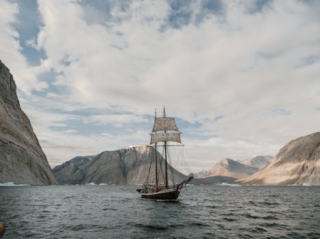 Majestic sailing ship traversing icy waters amidst grand rocky mountains under a cloudy sky.