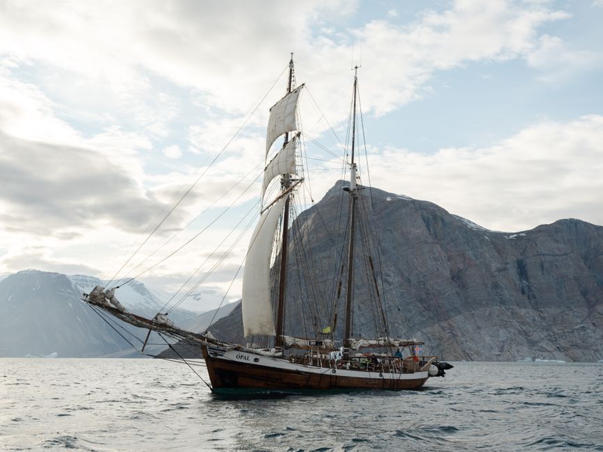 A classic schooner sails past dramatic rocky cliffs under a cloudy sky.