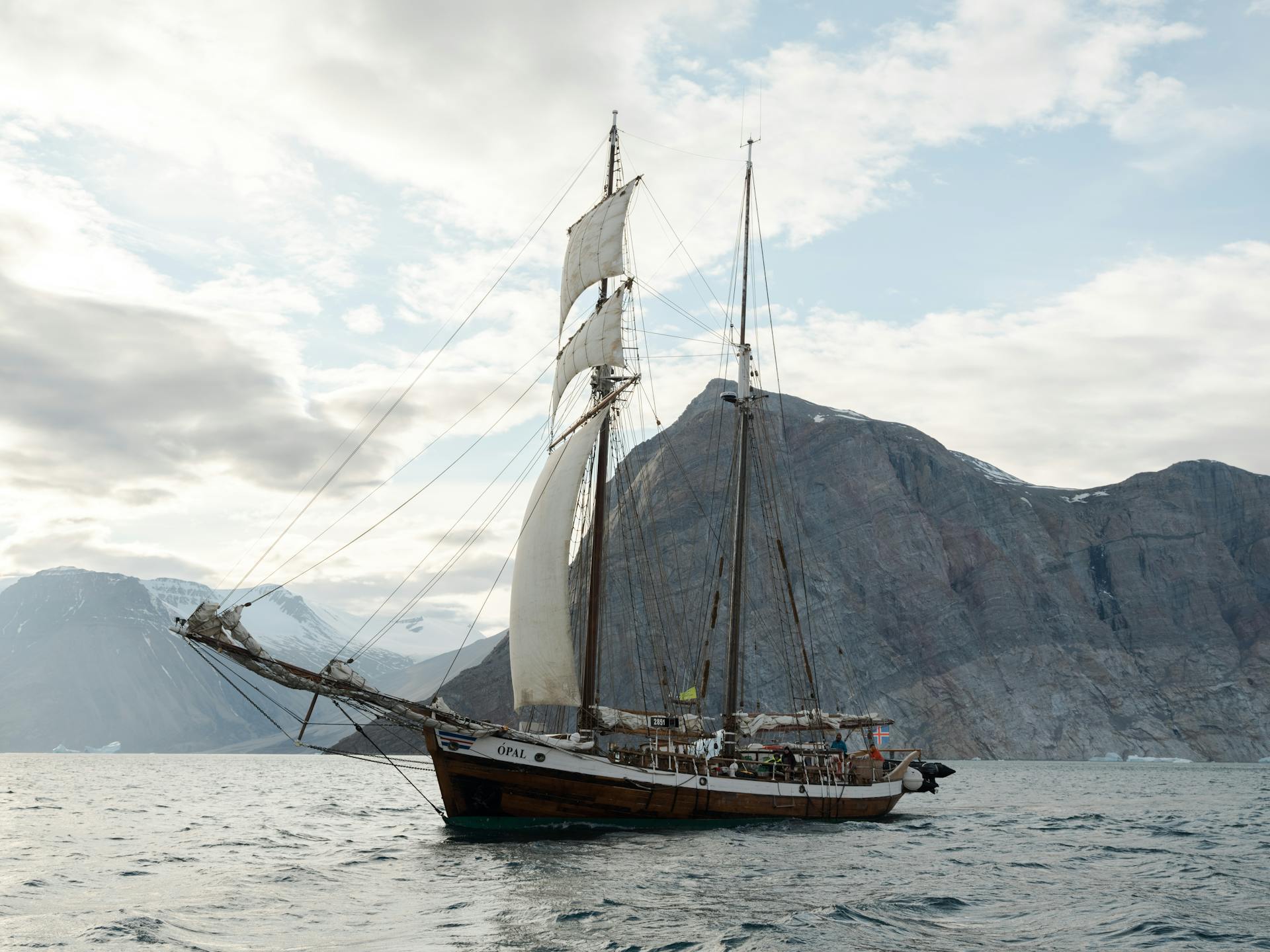 A classic schooner sails past dramatic rocky cliffs under a cloudy sky.