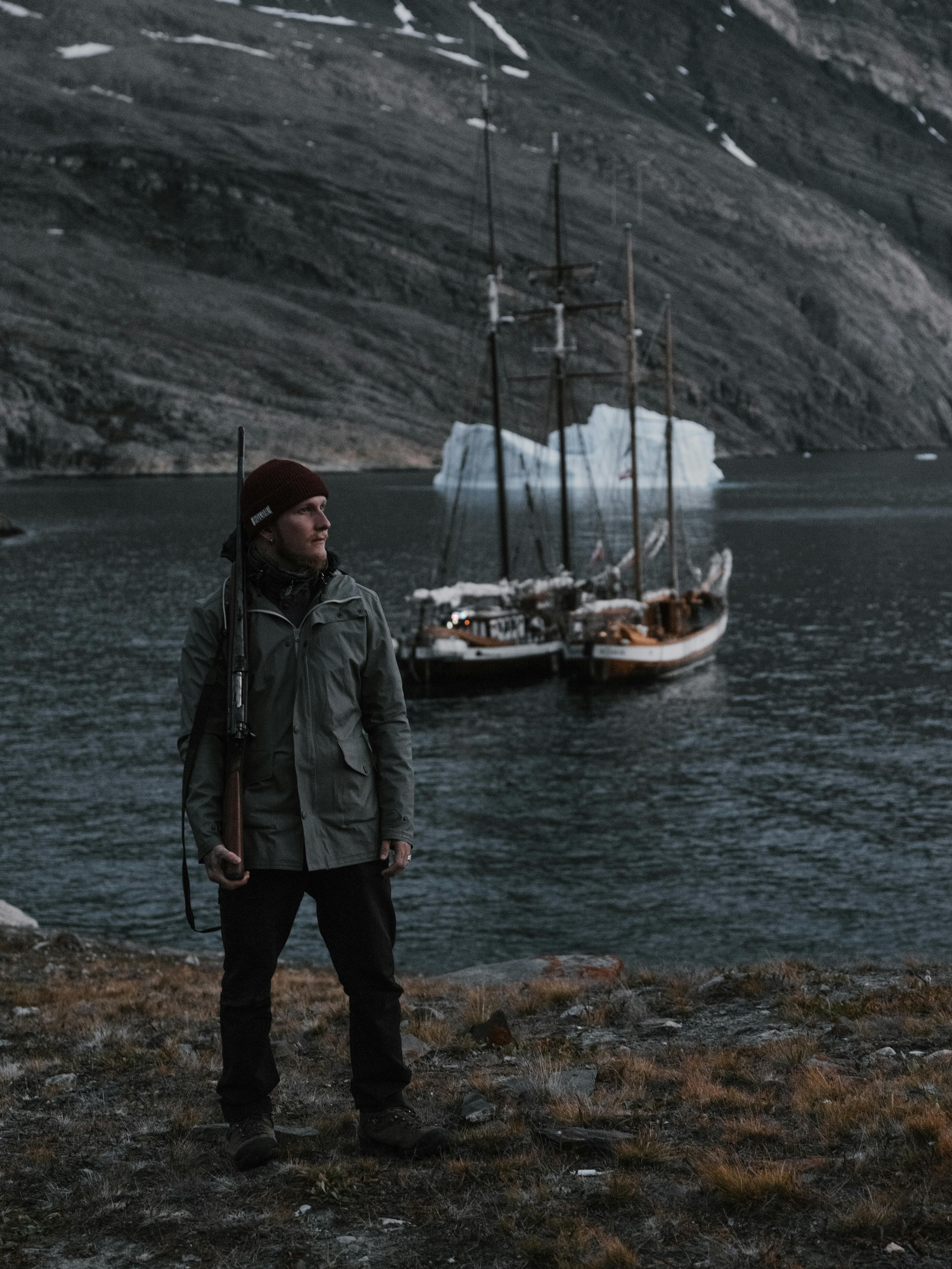 Portrait of a man with rifle by sailboats and iceberg in a remote fjord, showcasing rugged outdoor adventure.