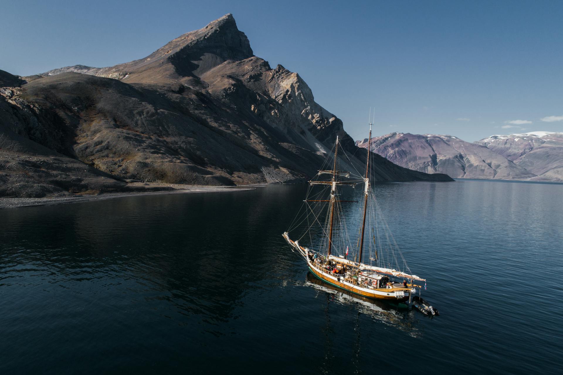 A schooner sails along a dramatic coastline with towering rock mountains under a clear sky.