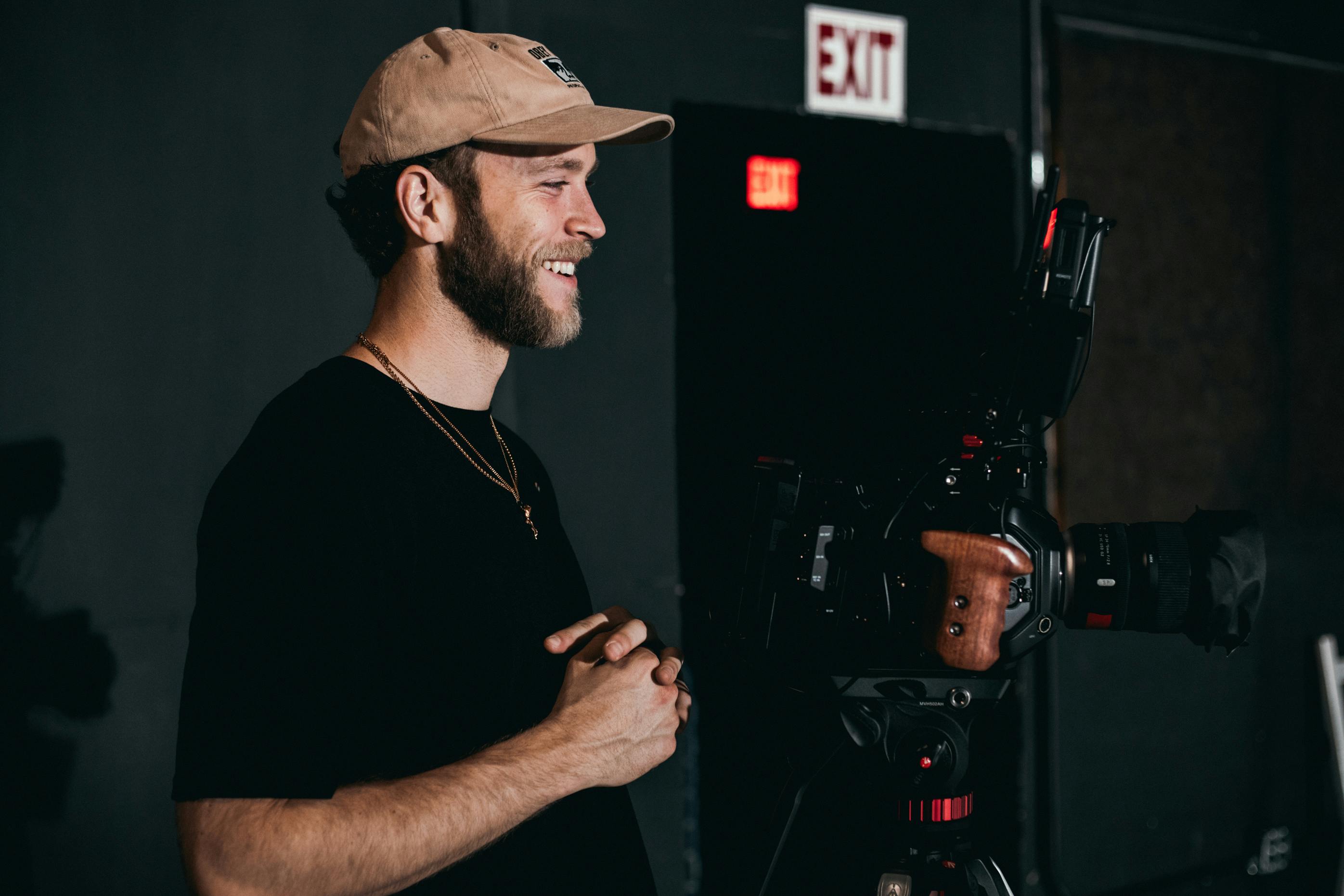 man in black crew neck t shirt wearing brown hat standing behind the camera on a tripod