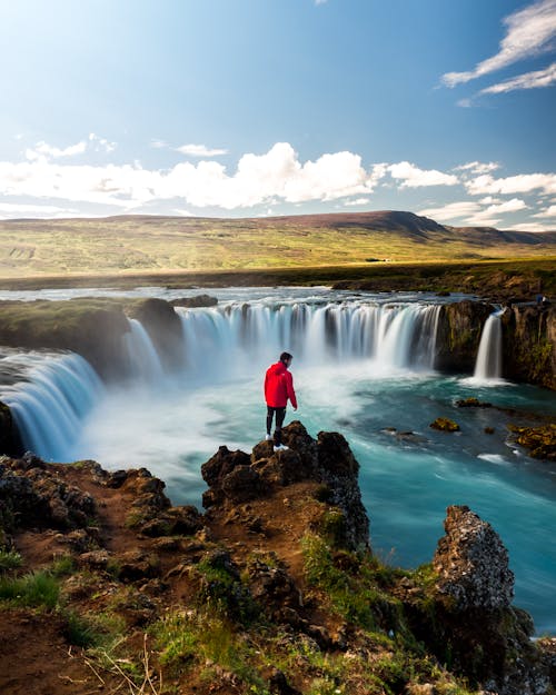 A Person in Red Jacket Standing on Brown Rock Near Waterfalls