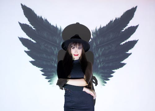 Pretty female with dark hair in hat standing with crossed arms under light near wall with projecting wings and looking at camera while casting shadow on background