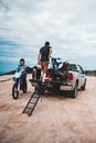 Unrecognizable male bikers loading motorbike in white pickup truck on sandy road in countryside against cloudy sky in nature outside