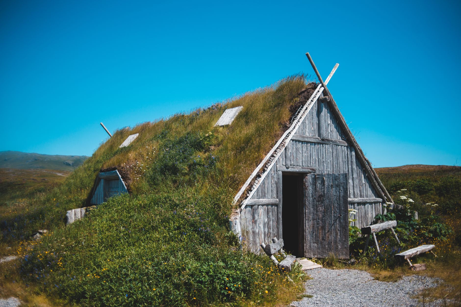 Exterior of shabby old turf roofed shed located on grassy terrain near hill against cloudless blue sky in Norstead Viking Village
