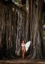 Full body of young slim female in swimsuit carrying surfboard and looking away near branchy tall tree