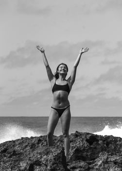Full body of cheerful glad female in swimsuit with eyes closed on rocky coast near waving ocean