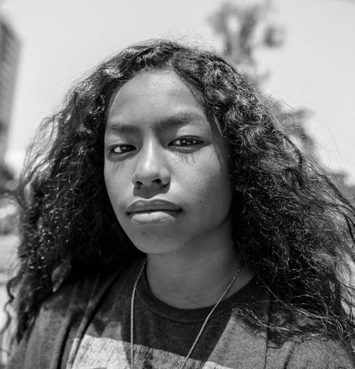 Black and white calm young female with curly hair wearing casual wear standing on blurred street on sunny summer day