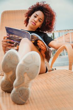 Low angle of trendy ethnic female with Afro hairstyle sitting with crossed legs on sun bed while reading magazine