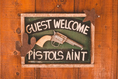 Framed rectangular signboard with inscription Guest Welcome Pistols Aint decorated with artificial gun and silhouettes of cactus and star on wooden door