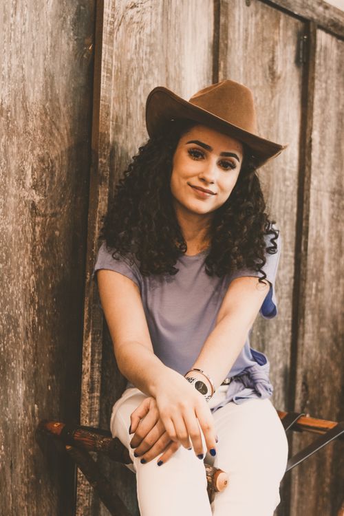 Free Charming young ethnic female with curly hair in hat looking at camera with half smile while relaxing in bright room with arms crossed on knees Stock Photo