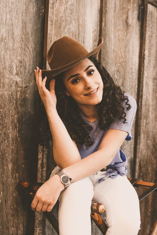 Free Beautiful young dark haired ethnic female in casual clothing touching hat and looking at camera with bright toothy smile while lounging on stool in bright room Stock Photo