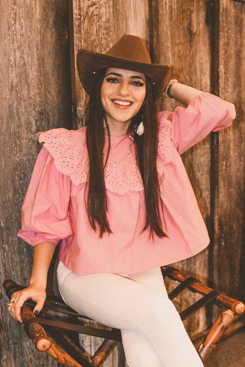 Cheerful young stylish female in pink blouse and white trousers touching hat and looking at camera with bright friendly smile