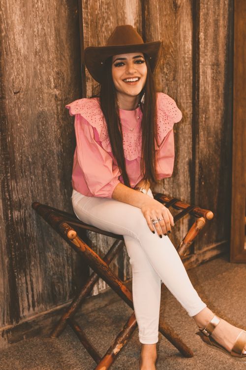 Free Full body of happy young slender female in stylish wear looking at camera with bright smile while relaxing on wooden camp chair in bright room in daytime Stock Photo