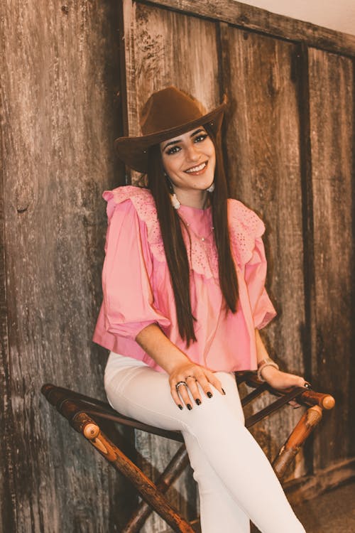 Charming young slender female in stylish pink shirt and white pants looking at camera with legs crossed and hand on knee while resting on folding chair by wooden wall