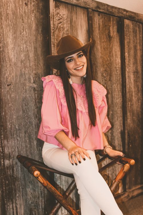 Free Charming young slender female in stylish pink shirt and white pants looking at camera with legs crossed and hand on knee while resting on folding chair by wooden wall Stock Photo
