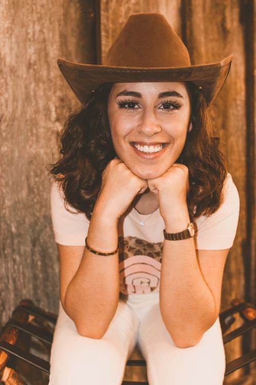 Free Cheerful young female in casual clothing and hat looking at camera with toothy smile with hands under chin Stock Photo