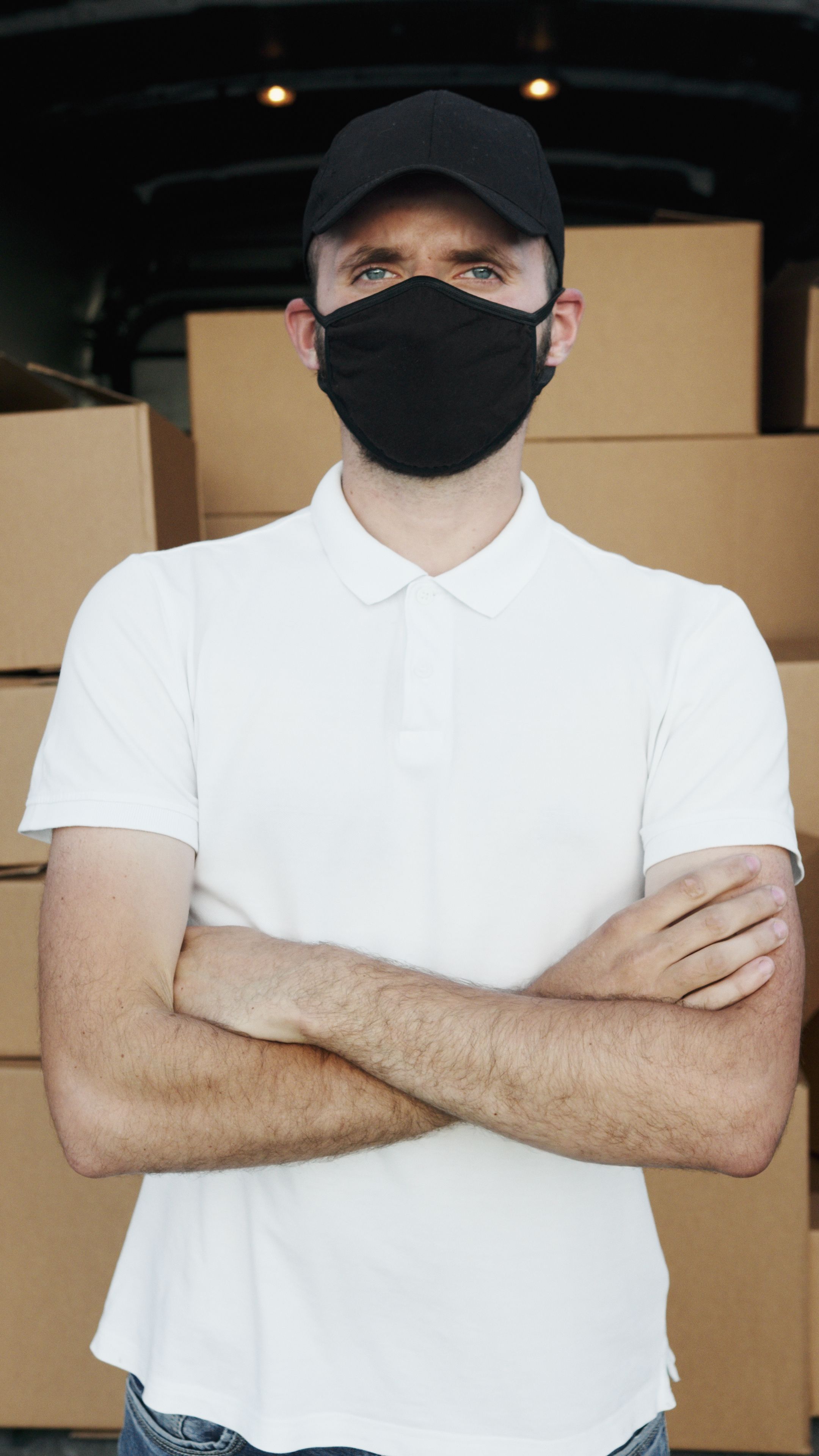 A courier in a white polo shirt, black mask, and hat standing with packages.